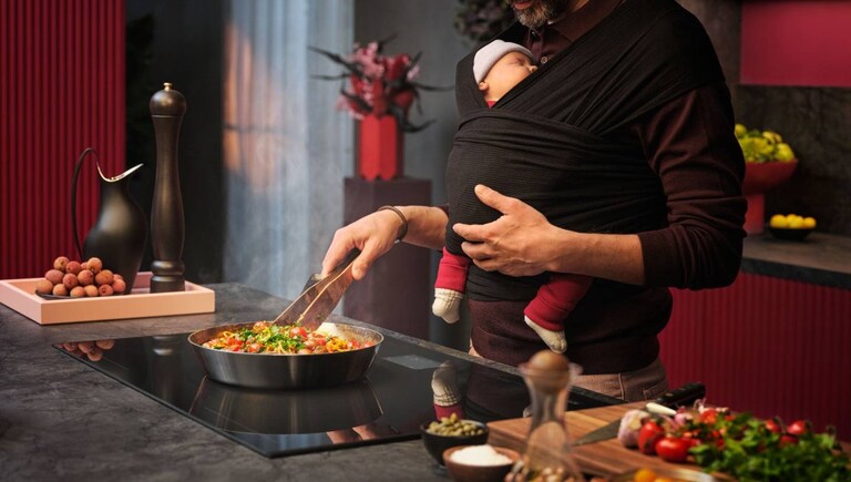 Man cooking food on a hob while carrying a baby in a baby carrier.